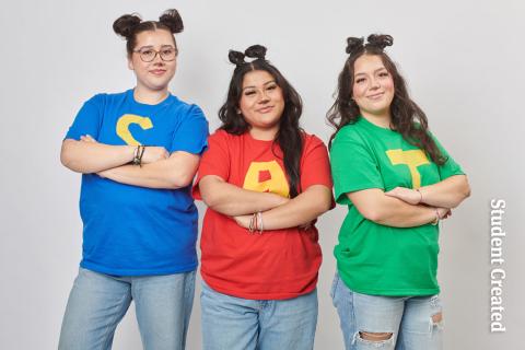 Student Photoshoot Fall 2024 Image of a student photoshoot with three female models. Their hair is done with two buns on top. the model on the left is wearing a blue shirt with an "S". the model in the middle is wearing a shirt with "A" on it and the third model on the right is wearing a green shirt with a "T" on it